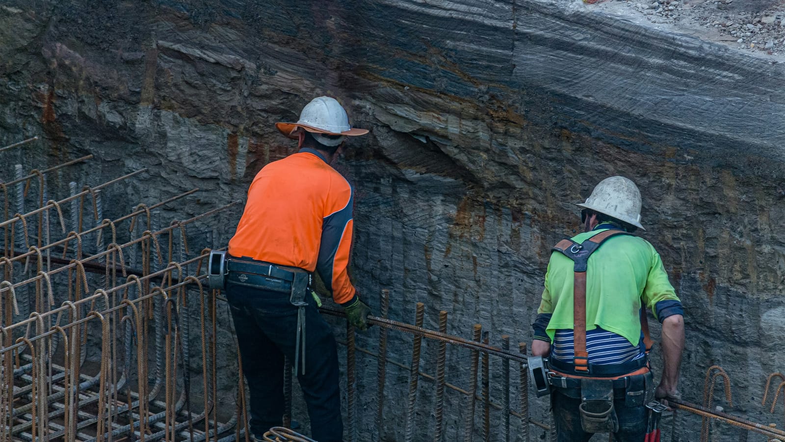 Two steel fixers in hi-vis shirts, hard hats and tool belts working on reinforcing bar at the foot of a rock-cut excavation, one wearing a fall-arrest harness
