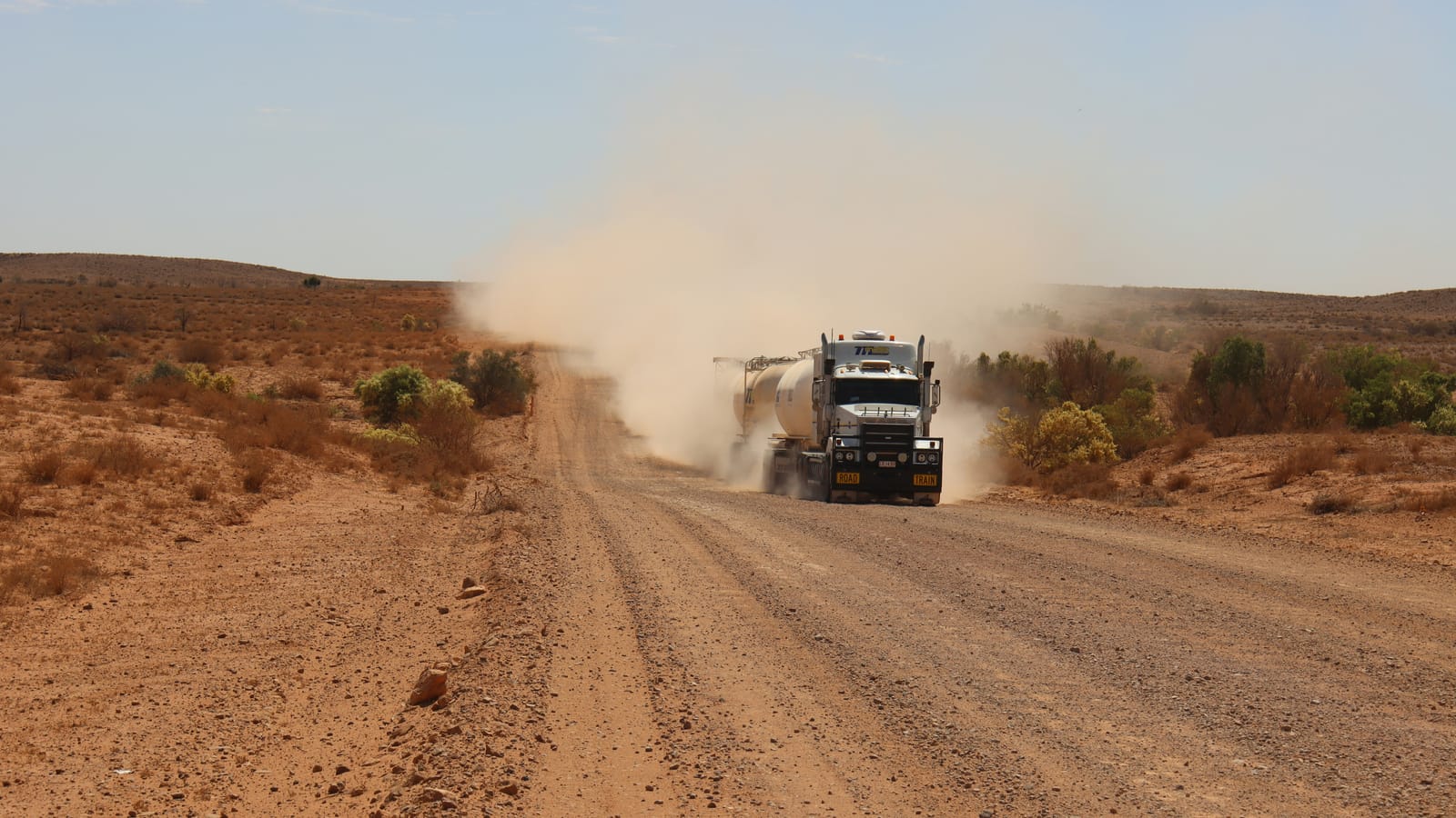 A road train tanker raising a cloud of dust on a gravel outback road, with a Road Train sign on its bullbar and low scrub on both sides