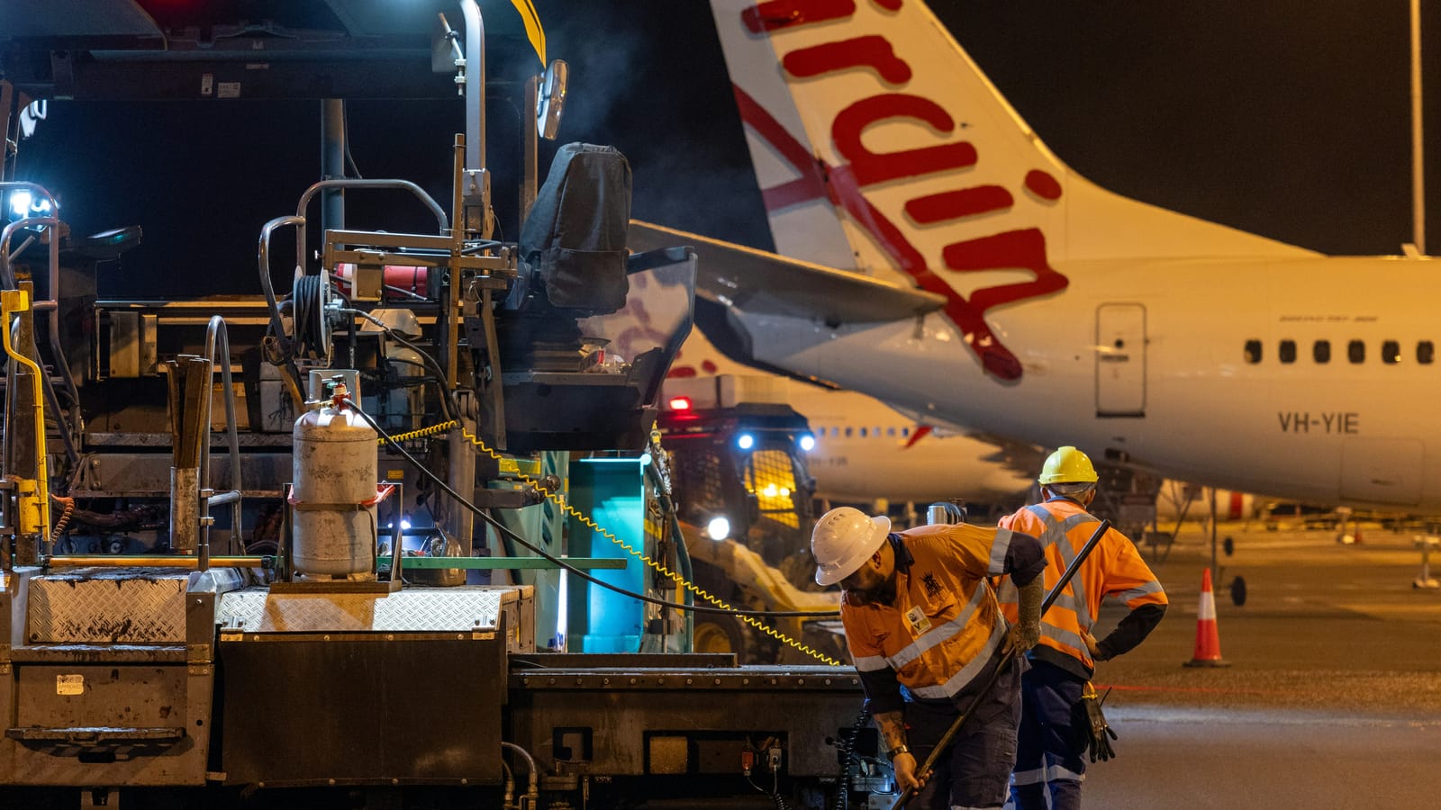 Night works crew in hi-vis operating an asphalt paver beside an aircraft on an Australian airport tarmac
