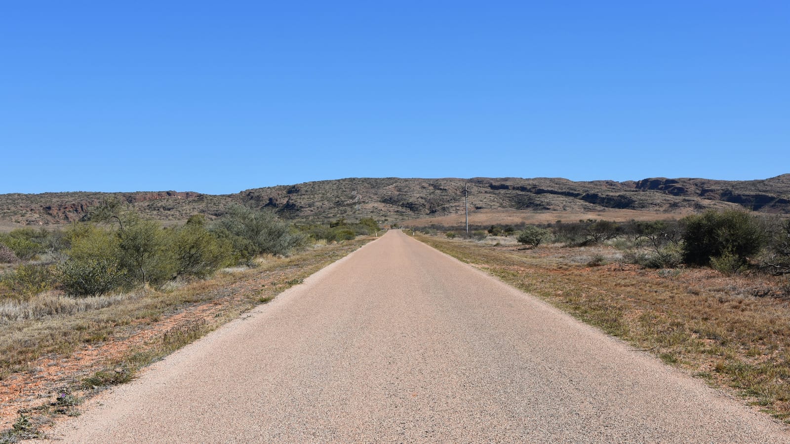 A sealed road running straight towards red ranges through outback scrub under a clear blue sky