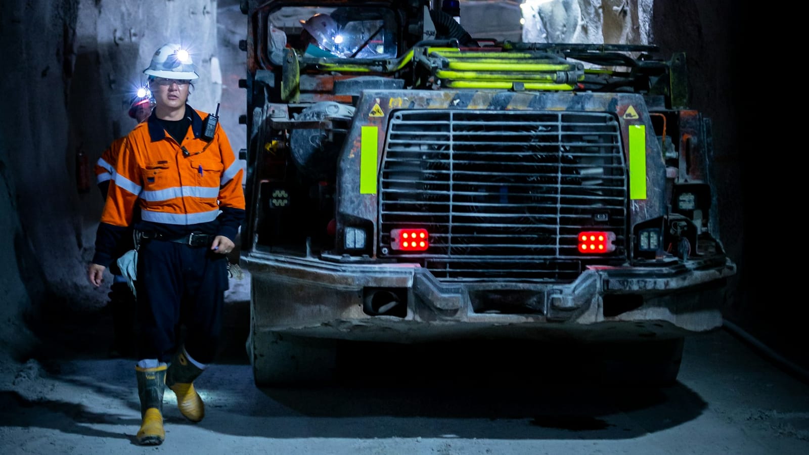 A worker in orange hi-vis with a cap lamp and radio walking past a loader in an underground mine drive at the start of a shift