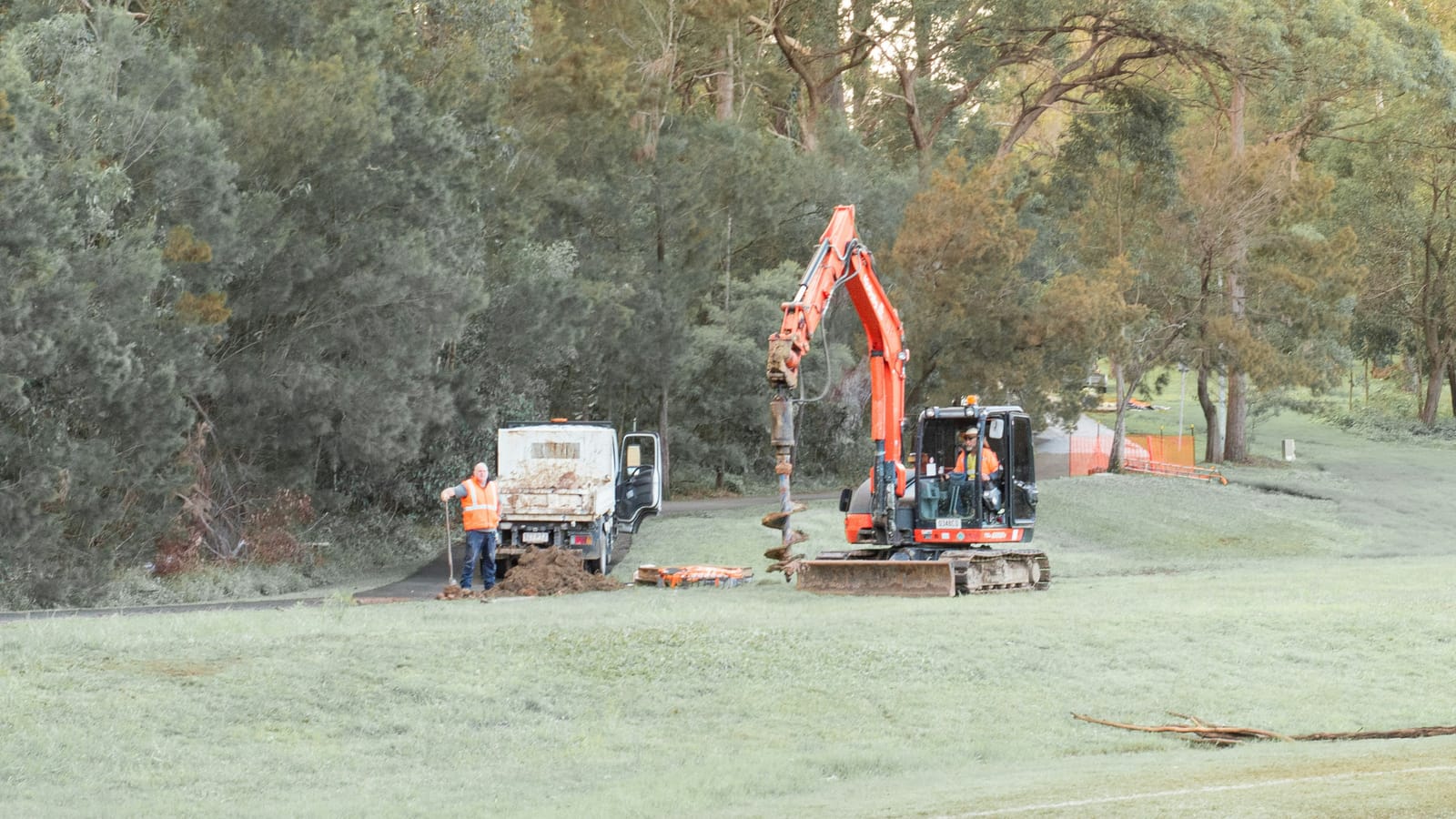 A small civil crew on a grassed reserve lined with gum trees, one worker in hi-vis beside a tipper truck while an operator runs a compact excavator fitted with an auger