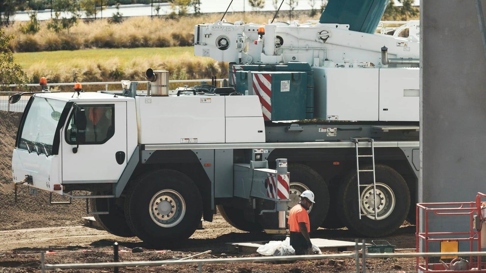 A worker in orange hi-vis setting an outrigger pad under a white all-terrain mobile crane on a dry site, with an EWP basket in the foreground