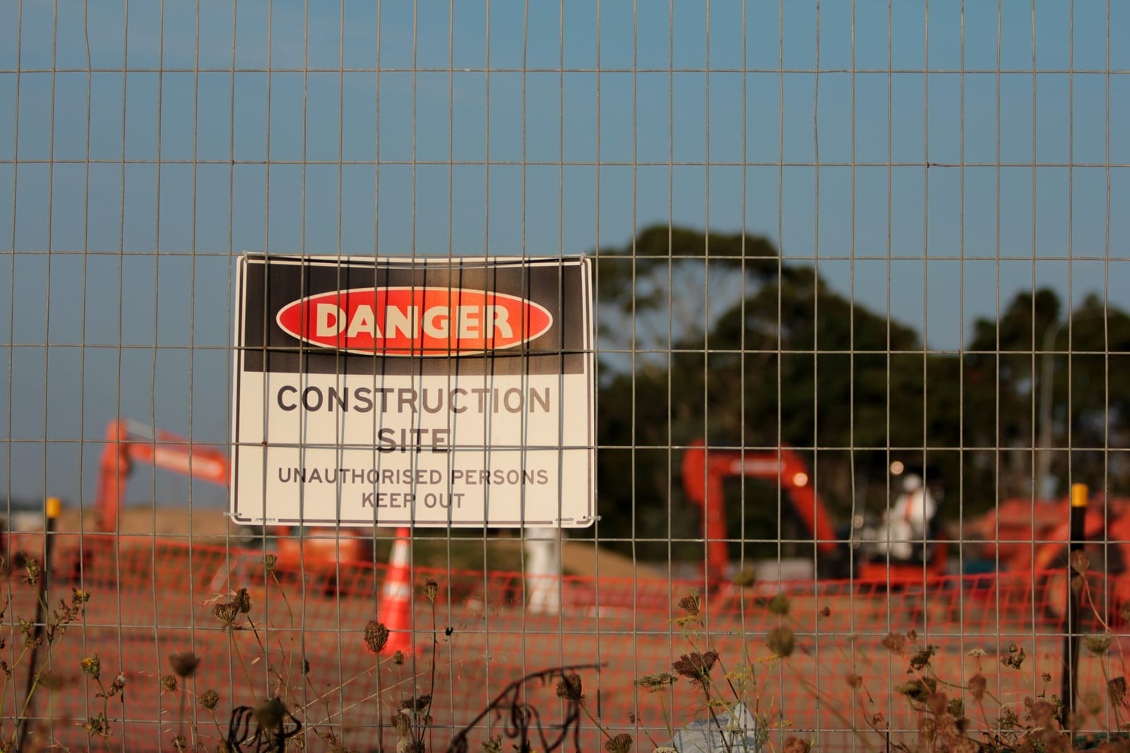 A danger sign reading construction site, unauthorised persons keep out, on temporary mesh fencing, with orange barrier mesh and excavators behind