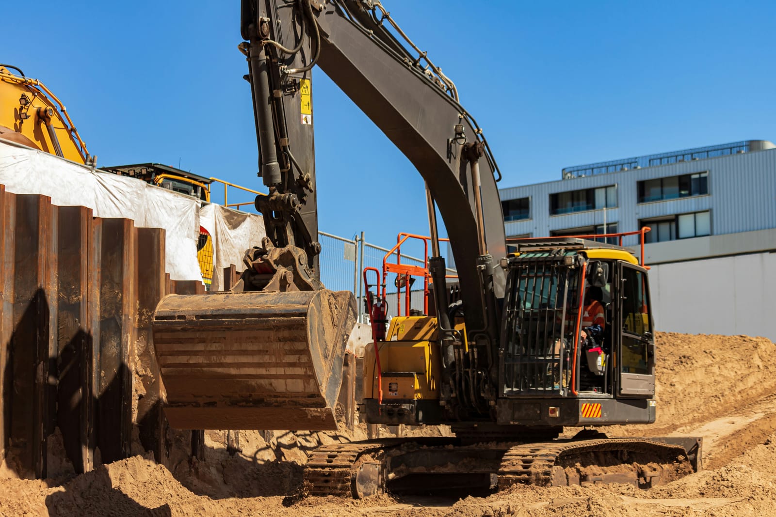 An excavator works a sandy excavation beside steel sheet piling, with temporary fencing and an elevated work platform behind