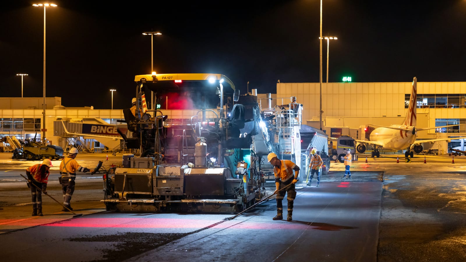 A night crew in hi-vis working around a paver on an Australian airport apron, the kind of blended on-hire workforce a host is responsible for