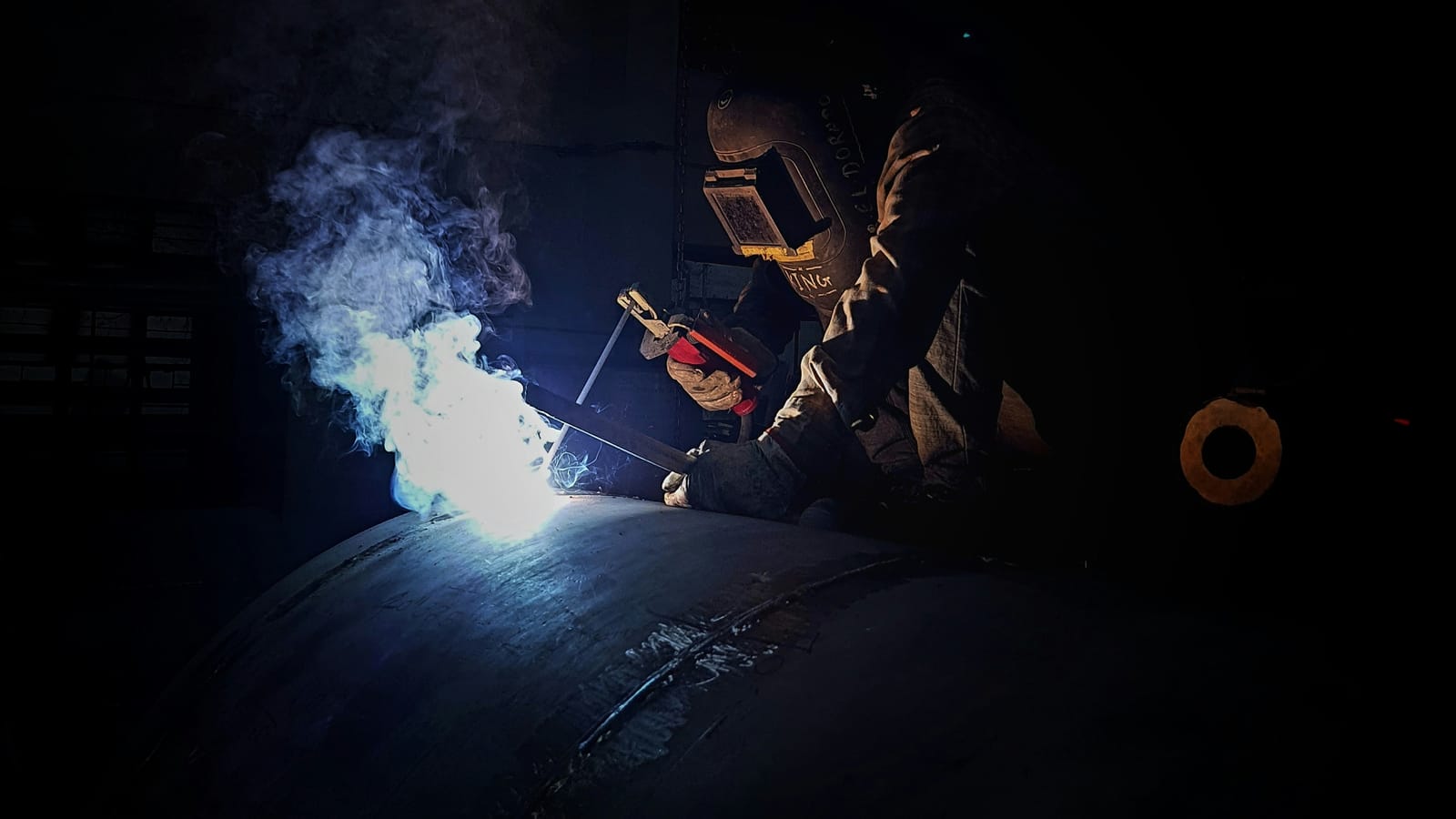 Welder working alone on a vessel seam in a darkened workshop, the night-shift end of a long roster