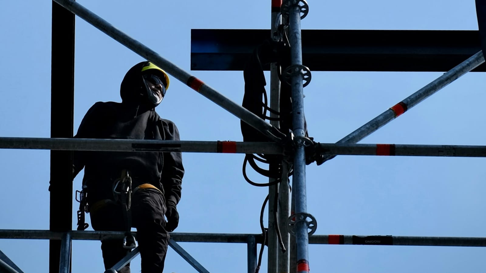 A rigger in a full-body harness and hard hat standing on a scaffold deck against a clear sky, lanyard and connectors at the hip
