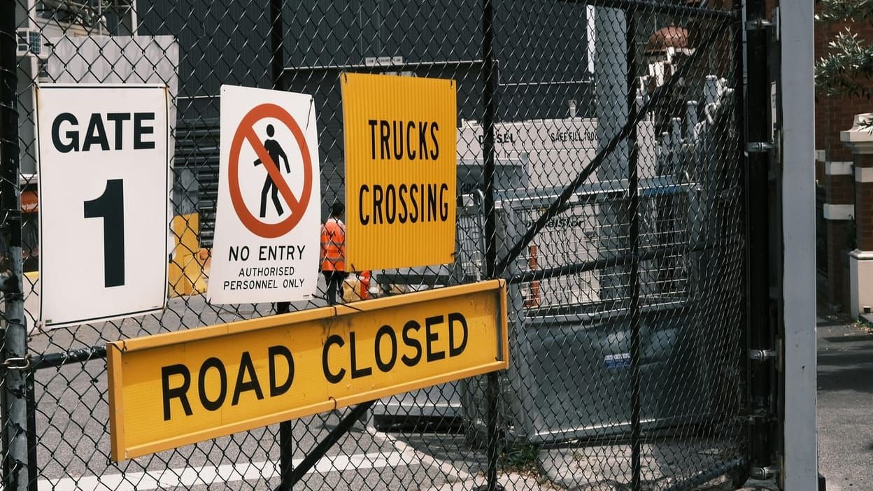 A locked construction site gate with Gate 1, no entry authorised personnel only, trucks crossing and road closed signs, and a worker in hi-vis behind the mesh