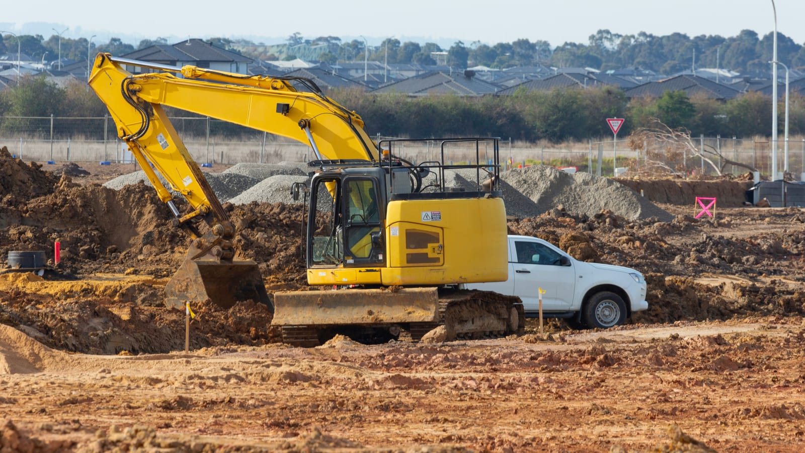 Yellow excavator digging beside a white work ute on a new housing estate civil site, with gravel stockpiles, temporary fencing and suburban rooftops behind