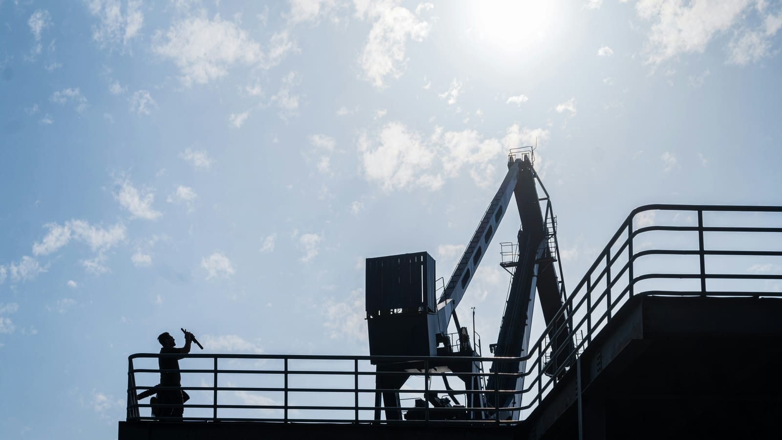 A worker silhouetted against a bright sky on the elevated walkway of industrial plant, crane boom overhead, the kind of structure a shutdown crew swarms over