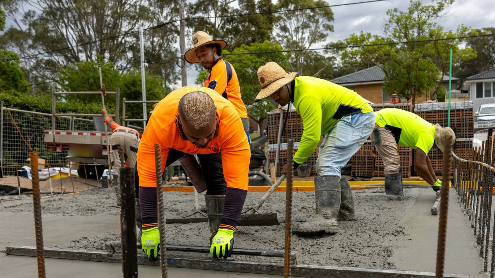 A concreting crew in orange and yellow hi-vis screeding a fresh slab between starter bars on a suburban Australian site