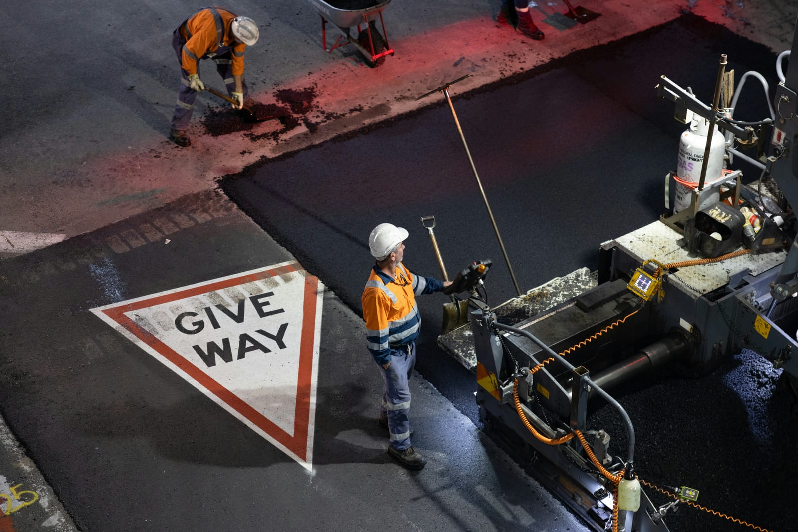 Two road workers in hi-vis and hard hats lay fresh asphalt at night beside a paver, next to a painted give way marking
