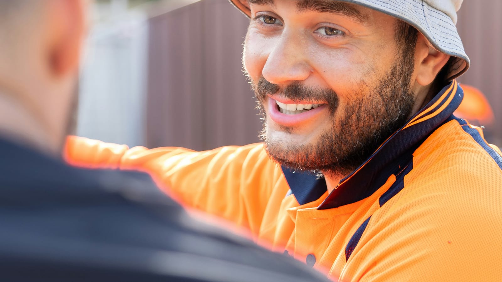 Two workers in orange hi-vis talking beside a site fence, one smiling mid-conversation