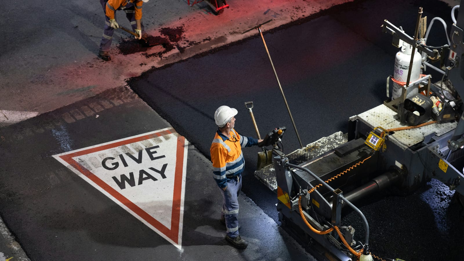 A road crew laying asphalt at night, with a paver operator in orange hi-vis and a white hard hat beside a freshly painted give way marking