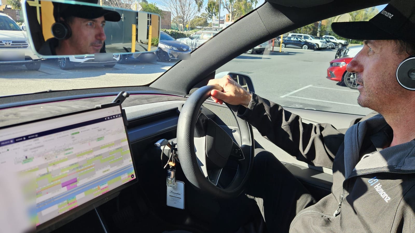 Duane Butler from Humanz in the driver's seat of a parked car in a Perth car park, headset on, one hand on the wheel, with a monitor mounted on the dashboard showing the Humanz scheduler board