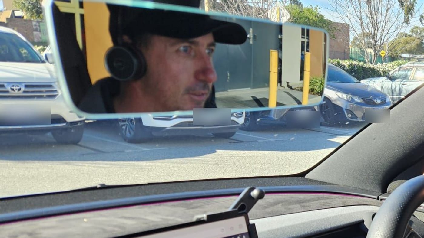 Duane Butler from Humanz reflected in the rear-view mirror of a parked car in a Perth car park, cap and headset on, with the dash monitor just in frame below. Car: parked.